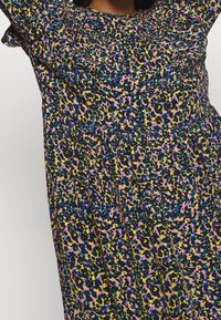 Femme portant une robe fleurie colorée avec un tissu froncé au niveau du col et de la taille, présentant des motifs roses, bleus, jaunes et noirs.