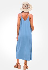 Himmelblaues, ärmelloses Maxikleid mit V-Ausschnitt und plissierter Textur, kombiniert mit einem Strohhut und braunen Blockabsatzsandalen.
