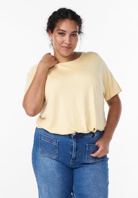 Cremefarbene Bluse mit kurzen Ärmeln und lockerem Schnitt, kombiniert mit blauen hoch taillierten Jeans mit zwei Gesäßtaschen. Weiche Textur.