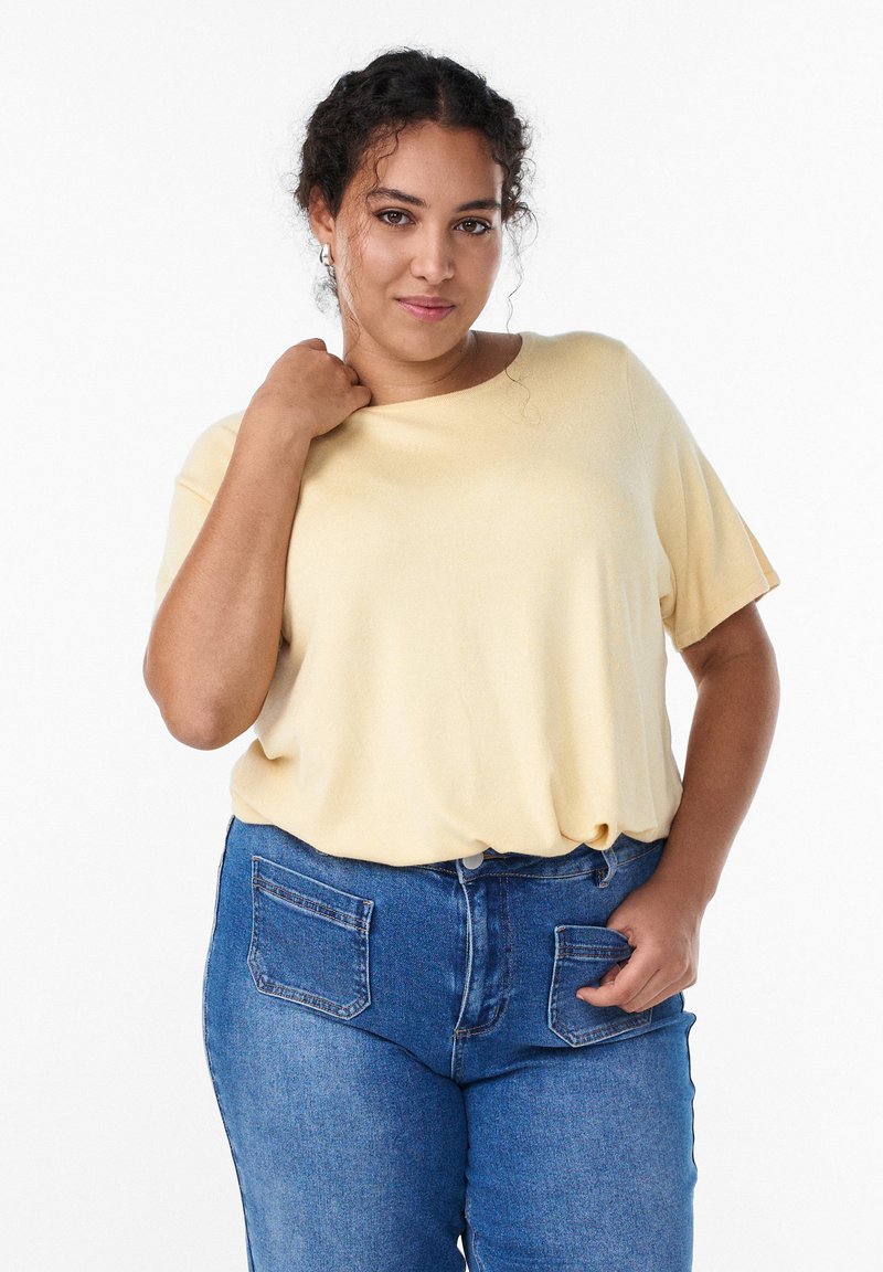 Cremefarbene Bluse mit kurzen Ärmeln und lockerem Schnitt, kombiniert mit blauen hoch taillierten Jeans mit zwei Gesäßtaschen. Weiche Textur.