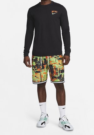 Homme portant un t-shirt noir à manches longues "Just Do It", un short graphique vert néon et orange, des chaussettes blanches Nike et des chaussures de sport blanches.