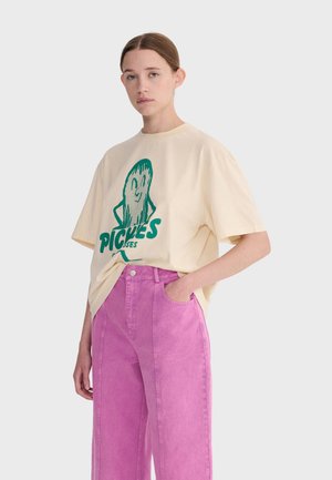 Mladá osoba s nízkym drdolom nosiaca nadrozmerné krémové tričko so zelenou karikatúrnou uhorkou a ružové široké nohavice, ruka v vrecku.