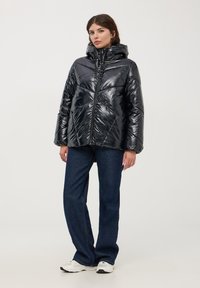 Giacca a vento nera con esterno lucido e trapuntato, cappuccio aderente e chiusura con zip, abbinata a jeans blu e sneaker.