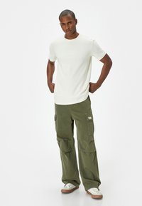 Ljusbeige kortärmad T-shirt, gröna cargobyxor med sidofickor och vita sneakers, stående framför en vit bakgrund.