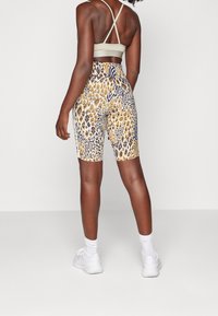 Högmidjade shorts med ett guld- och blått djurmönster, tillverkade av stretchigt tyg. En ljusfärgad sport-bh bärs ovanpå. Vita skor syns.