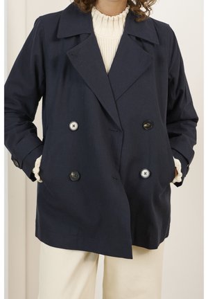 Femme portant un manteau bleu marine croisé sur un pull en maille crème, les mains dans les poches du manteau, avec un pantalon beige visible.