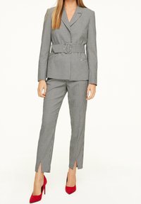 Un costume à motif pied-de-poule avec une ceinture, comprenant un blazer ajusté et un pantalon droit. Des chaussures à talons hauts rouges complètent le look.