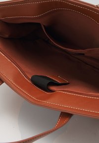 Sac fourre-tout synthétique marron avec des coutures blanches, présentant un intérieur ouvert, une poche zippée et un attachement de sangle noir.