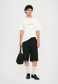 T-shirt bianco oversize con grafica gialla, pantaloni corti neri fino al ginocchio, scarpe nere slip-on e calzini grigi. Borsa nera tenuta nella mano sinistra.