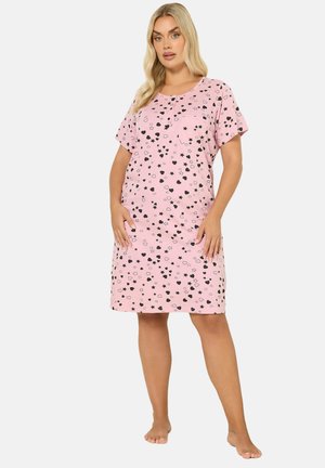 Frau trägt ein rosa knieumspieltes Kleid mit kurzen Ärmeln und schwarzen und weißen Stern- und Herzmustern, steht barfuß auf weißem Hintergrund.