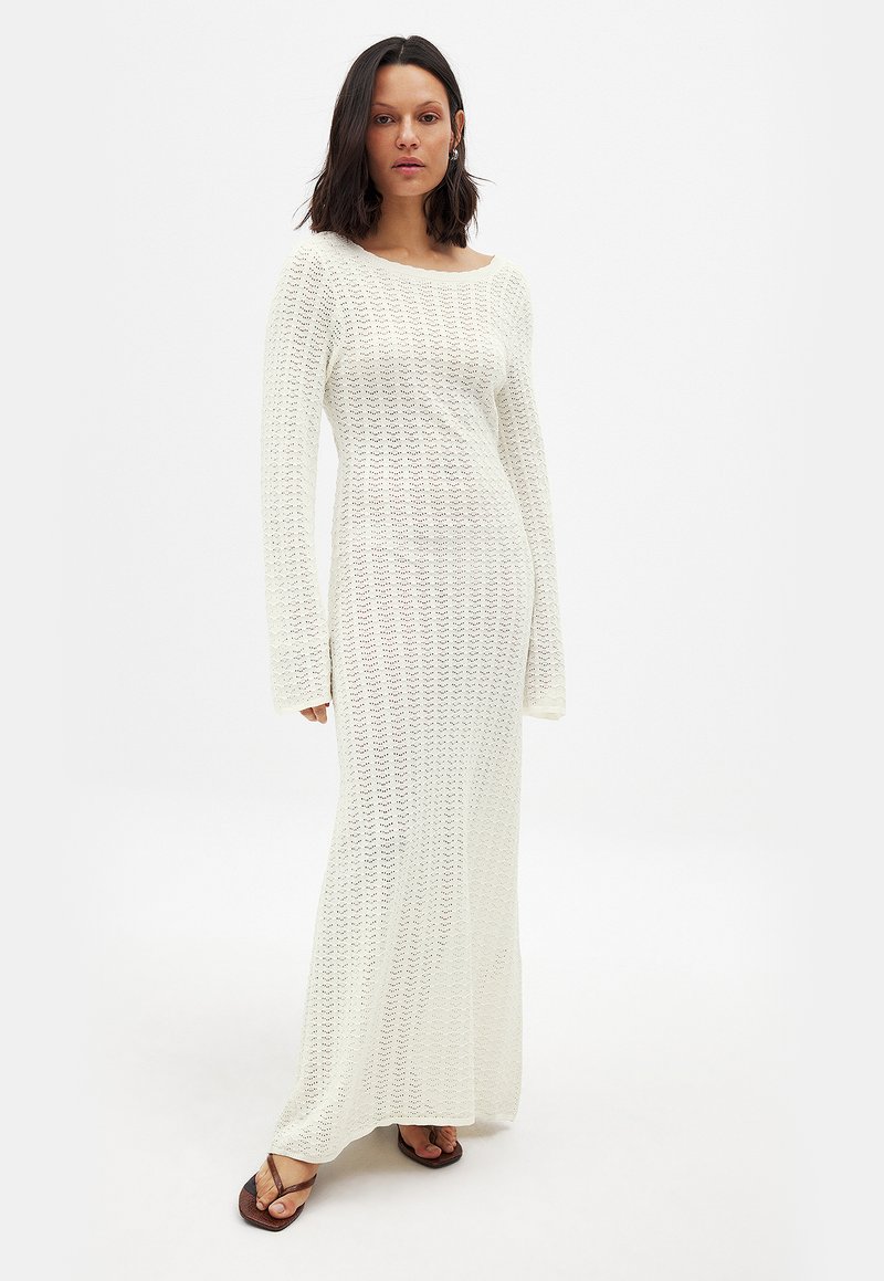 Langes, weißes Strickkleid mit einem Bootsausschnitt, langen Ärmeln und einem offenen Strickmuster. Kombiniert mit braunen Sandalen. Weiche Textur, bodenlang.