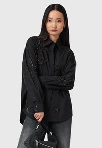 Camicia nera con bottoni, decorata con perline, dotata di maniche lunghe, colletto e vestibilità rilassata. Indossata con jeans grigi e una borsa nera.