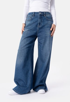 Kvinde iført blå jeans med vide ben og hvide sneakers, står med afslappede hænder ned langs siderne mod en ensfarvet baggrund.