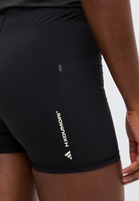 Κοντινό πλάνο ατόμου που φοράει μαύρο αθλητικό σορτς με το λογότυπο "H.KOU.MORI" και το σύμβολο Adidas στον αριστερό μηρό.