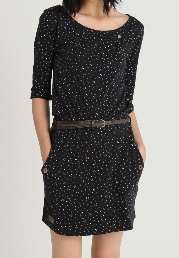 Femme portant une robe noire avec de petits motifs floraux roses et blancs, des manches trois-quarts, deux poches à boutons, et une ceinture tressée marron.