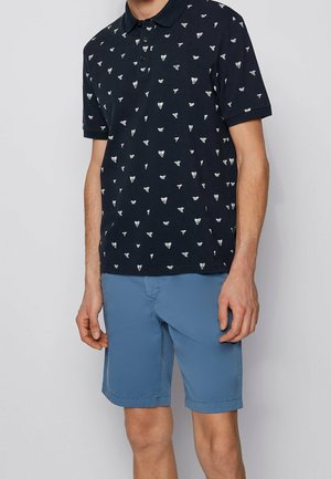 Homme portant un polo bleu marine avec des motifs abstraits blancs et un short bleu clair arrivant aux genoux, sur un fond uni.
