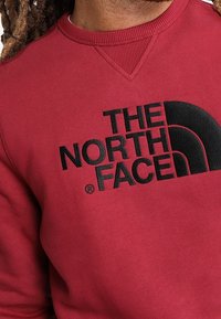 Sudadera roja con cuello redondo, que presenta un gran logo negro "THE NORTH FACE". Textura de tela suave con detalles de cuello acanalado.