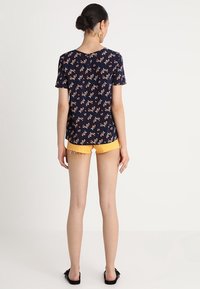 Tmavě modrý top s krátkým rukávem s květinovým vzorem v broskvové a bílé barvě, doplněný žlutými šortkami. Hladká textura a volný střih.