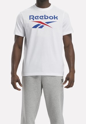 Mann trägt weißes Reebok-T-Shirt mit blau-rotem Logo und graue Reebok-Sweathose, steht vor einfarbigem Hintergrund.