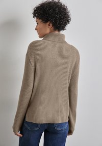 Beige svetr s ribovaným vzorem, vysokým výstřihem a dlouhými rukávy, s volným střihem a bočními rozparky, spárovaný s modrými džíny.