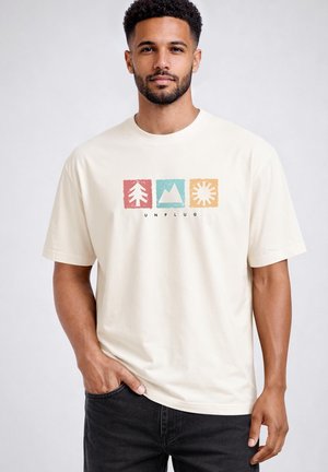 Man wearing a cream T-shirt with red, blue, and yellow blocks showing a tree, mountain, sun, and the word "UNPLUG" on the front.