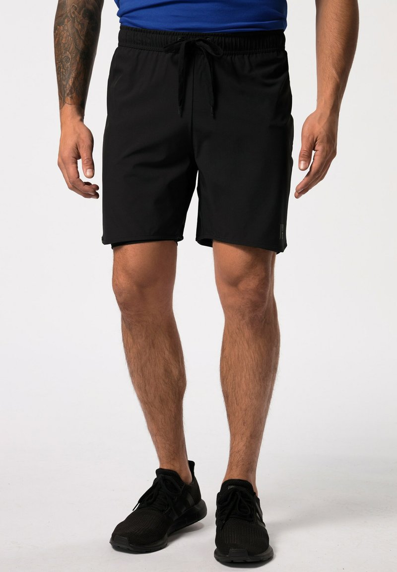 Schwarze sportliche Shorts mit einem Kordelzugbund, glatter Textur, in mittlerer Oberschenkellänge und subtilen Seitenpaneelen. Getragen mit schwarzen Turnschuhen.