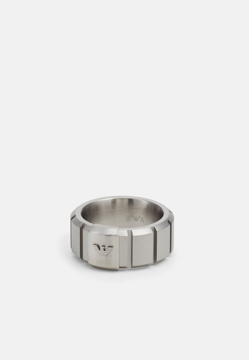 Metalring med en poleret sølvfinish. Har et bredt design, geometriske kanter og et indgraveret logo på en flad overflade.