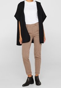 Kobieta w beżowych spodniaczkach w prążki, białej koszulce, czarnym bezrękawniku z otwartym przodem i czarnych botkach sięgających kostek, stojąca na tle jednolitym.