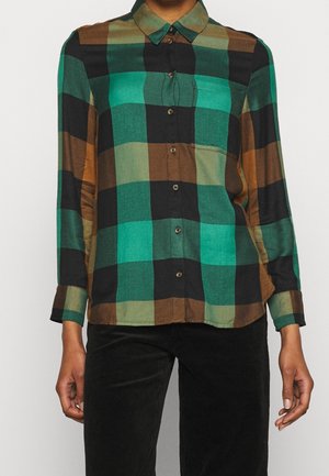 Femme portant une chemise à manches longues avec un grand motif à carreaux verts, noirs et marron, et un pantalon noir.