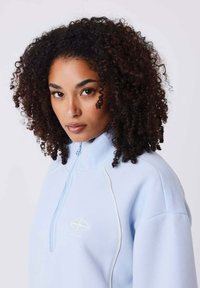 Sweat à col zippé bleu clair fabriqué en tissu doux, présentant un logo et des coutures blanches contrastantes, avec un col haut et une coupe décontractée.