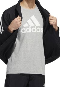 Svart idrottsjacka med vita ränder, lager på lager över en ljusgrå t-shirt med en framträdande vit adidas-logotyp. Texturerad interiör synlig.