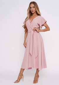 Rosa Wickelkleid mit schwingenden Ärmeln, tailliertem Gürtel und fließender Midi-Länge. Kombiniert mit hautfarbenen, gestrickten Absatzschuhen. Glatte Stoffoberfläche.