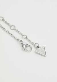 Accesorio de cadena de plata con un colgante triangular con el logotipo "GUESS" en relieve y un seguro cierre de langosta. Textura de metal suave.