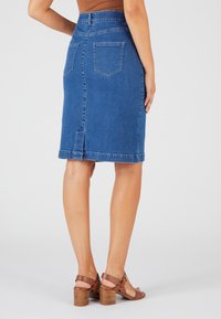 Jupe en denim bleu foncé, à mi-longueur, avec une coupe droite, des poches arrière et une fente au bas. Associée à des sandales à talons bruns.