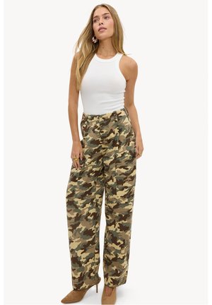 Femme aux longs cheveux blonds portant un haut blanc sans manches, un pantalon camouflage taille haute et des chaussures à talons hauts couleur beige, debout.