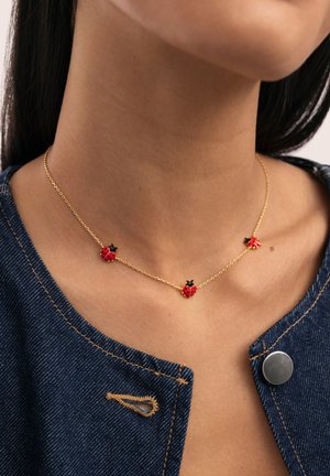 Collar de cadena dorada con tres pequeños colgantes de mariquitas rojas, llevado en el cuello de una persona, combinado con una prenda de mezclilla azul.