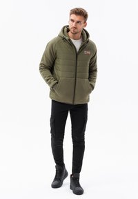 Felpa con cappuccio imbottita verde oliva con zip e cordoncino, caratterizzata da un patch con logo, abbinata a pantaloni cargo neri e sneakers alte nere.