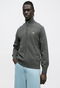 Sivý sveter na zips s rebrovaným golierom a manžetami, vyrobený z mäkkého materiálu, s malým zeleným logom na ľavej strane.
