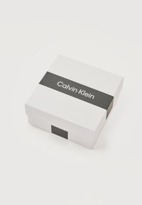 Λευκό τετράγωνο κουτί με μαύρη οριζόντια ταινία που φέρει το λογότυπο "Calvin Klein" σε λευκό φόντο.