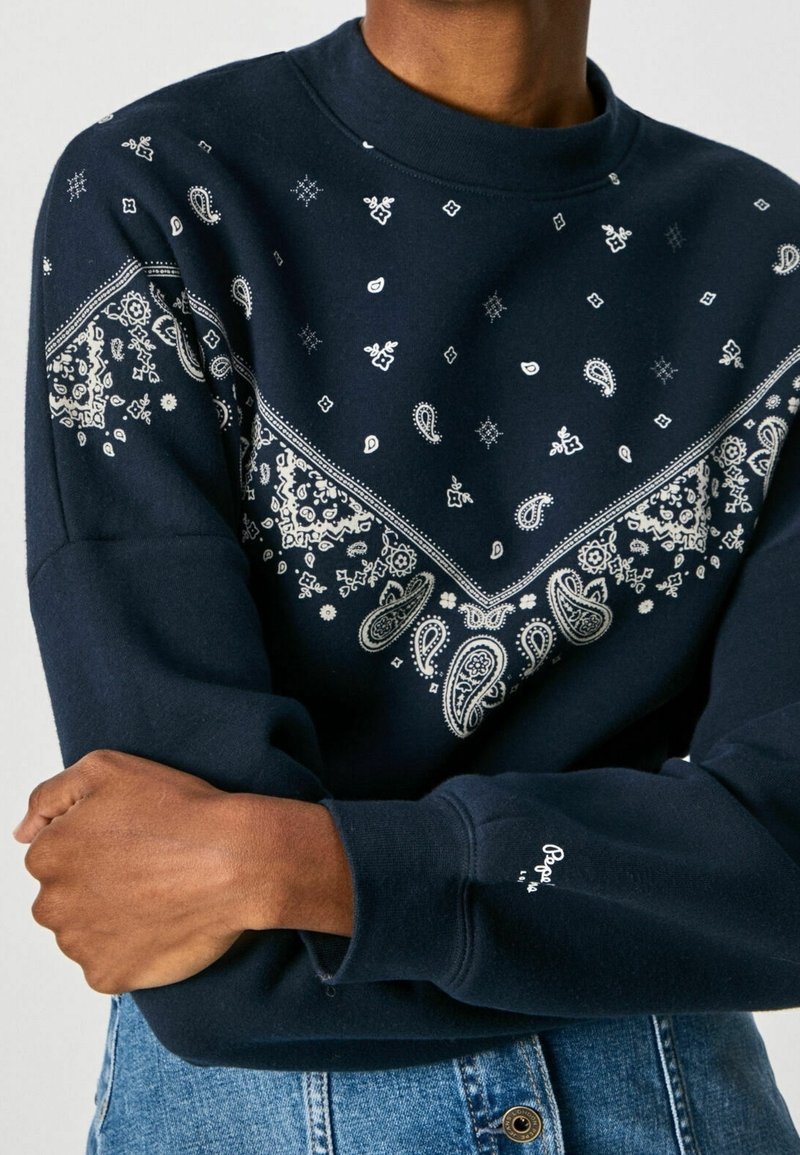 Sweat-shirt bleu marine avec motifs paisley blancs et style bandana, porté par une personne les bras croisés sur un jean en denim bleu.