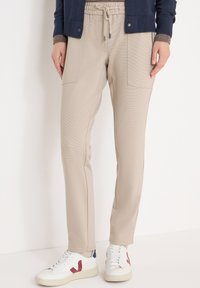Beige sweatpants met twee steekzakken aan de voorkant en een elastische tailleband, gecombineerd met witte sneakers met rode en blauwe accenten.
