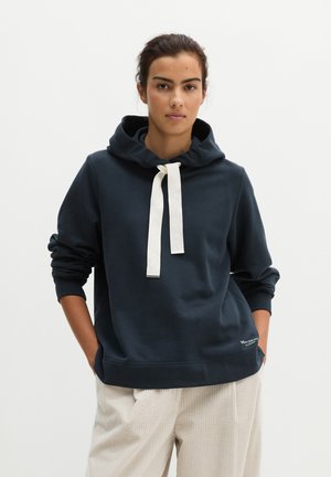 Femme portant un sweat à capuche bleu foncé avec de larges cordons blancs et un pantalon beige clair, debout les mains dans les poches devant un fond uni.