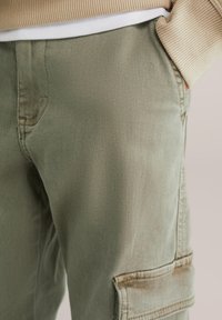 Pantalones cargo en verde oliva claro, hechos de una suave mezcla de algodón, con bolsillos laterales y una textura ligeramente desgastada para un estilo casual.