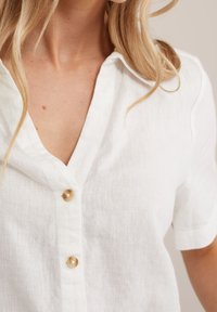 Vit linne skjorta med V-hals och korta ärmar, med tre beigefärgade knappar framtill och en strukturerad tyg.