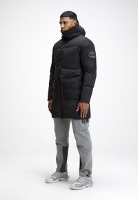 Giacca a vento nera con colletto alto e chiusura a zip. Presenta una toppa con logo sulla manica, indossata sopra pantaloni sportivi grigi e sneakers.