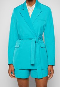 Blazer turchese con cintura legata, revers a scollatura, e taschino sul petto, abbinato a pantaloni corti coordinati. Tessuto morbido e vestibilità sartoriale.