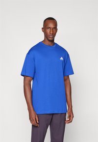 Blue cotton t-shirt with a crew neck, short sleeves, and a white Adidas logo on the left chest. Paired with dark pants.