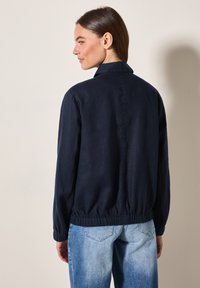 Donkerv blauwe jas met een kraag, elastische tailleband en lange mouwen, gemaakt van lichte stof, gecombineerd met lichtblauwe denim jeans.