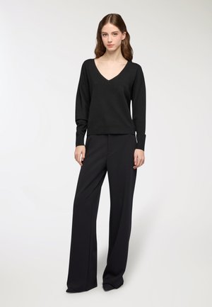 Donna in piedi che indossa un maglione nero a maniche lunghe con scollatura a V e pantaloni neri larghi, su uno sfondo bianco semplice.