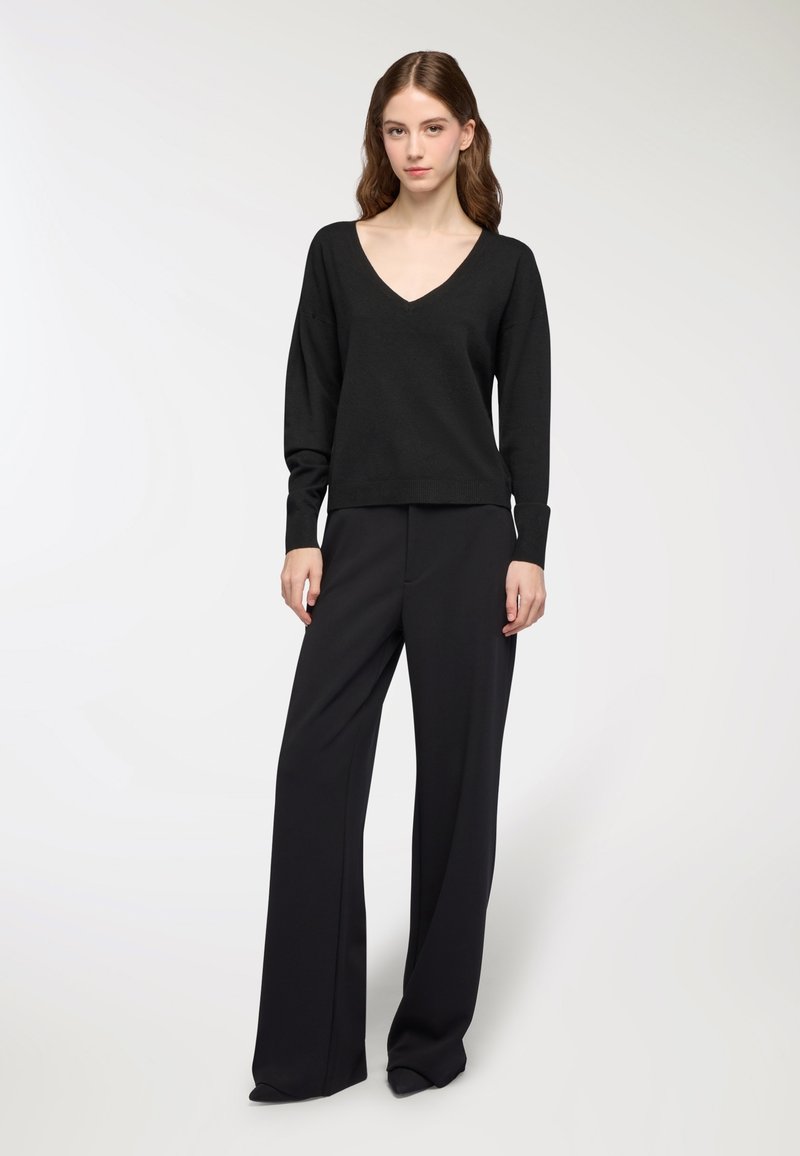 Kobieta stojąca w czarnym swetrze z dekoltem w serek i szerokich czarnych spodniach na tle jednolitego białego tła.